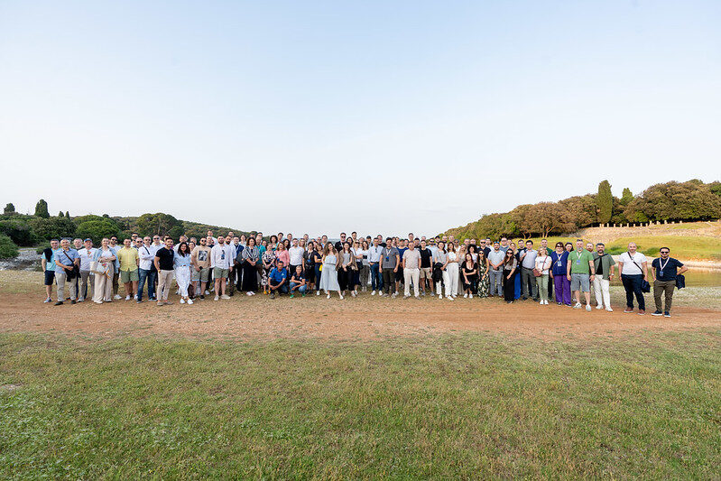 The entire conference delegation, well over a hundred people, standing together in an open meadow in evening light.