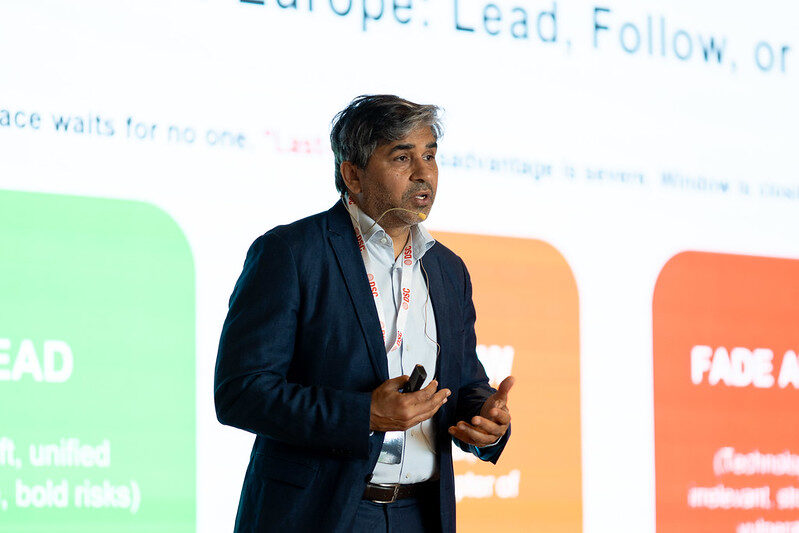 Tarry Singh speaking with a headset microphone in front of a slide reading Europe: Lead, Follow, or Fade Away.
