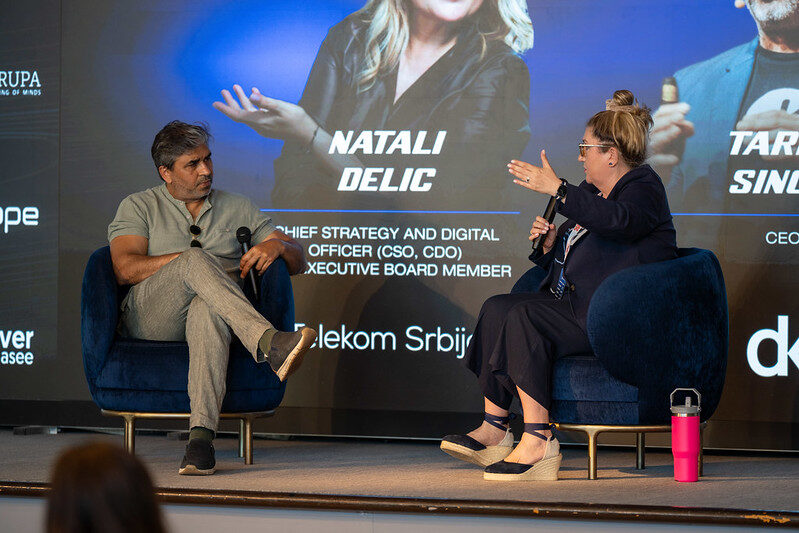 Tarry Singh and Natali Delic seated in armchairs on stage during the fireside chat, their names and titles projected behind them.