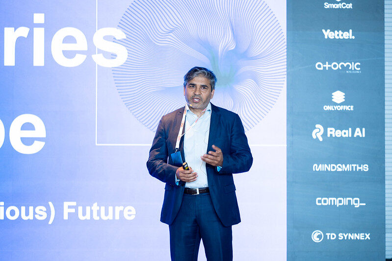 Tarry Singh mid-sentence on stage, partner logos on the rail to his side.