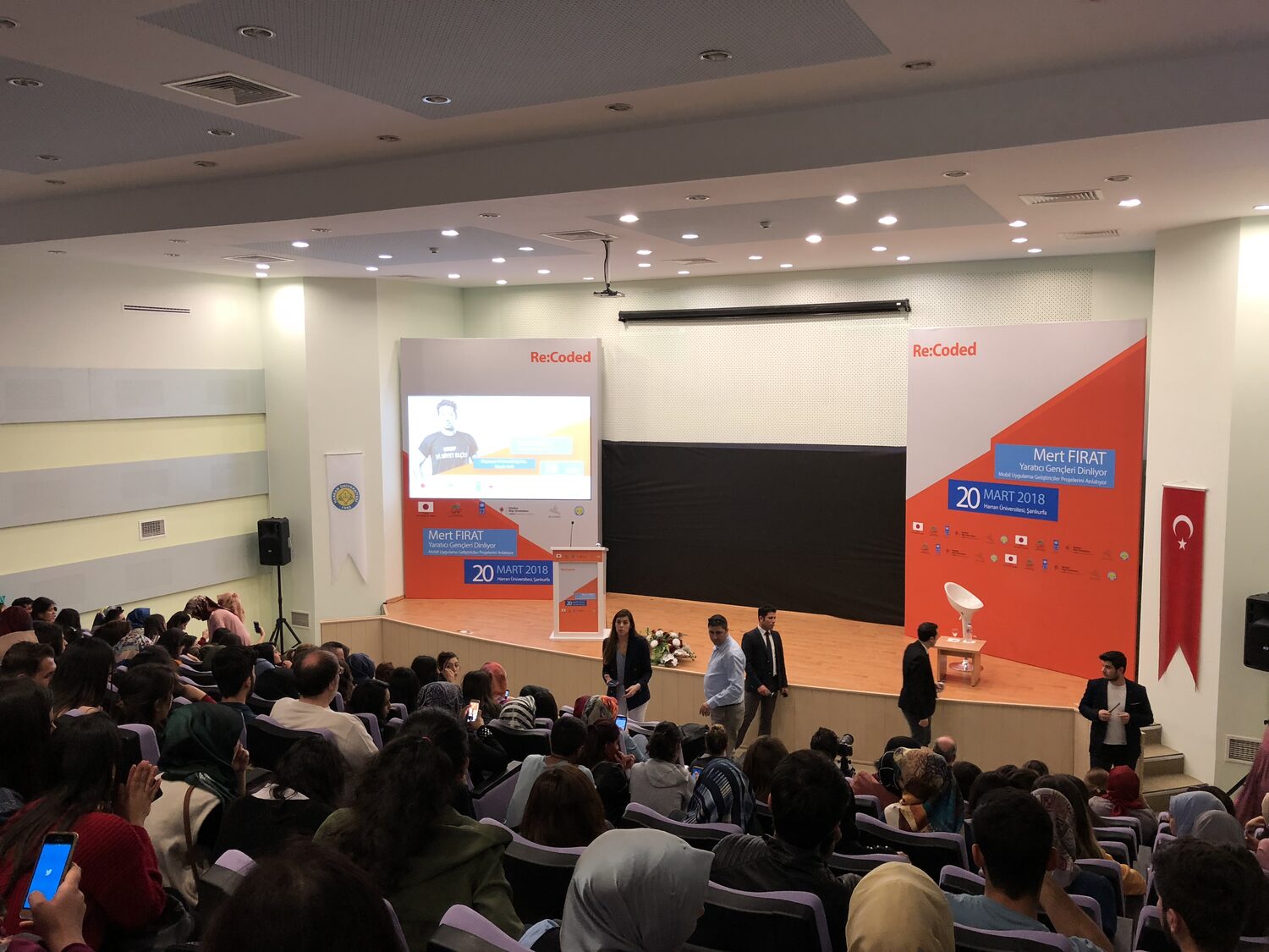 A packed auditorium at Harran University, Şanlıurfa, facing a stage with Re:coded boards printed 20 Mart 2018