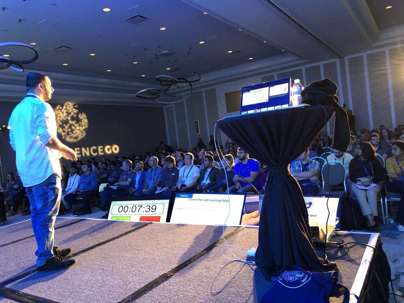 Tarry Singh speaking on the DataScienceGO main stage in San Diego, mid-gesture in front of a full ballroom, a slide reading Current Pain With Learning Rates on the screen behind him and a countdown clock showing seven minutes remaining.