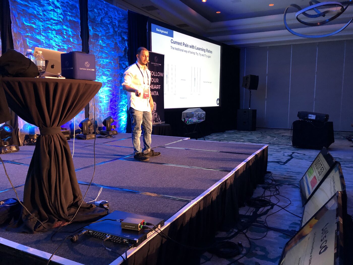 Wide view of the DataScienceGO main stage, Tarry Singh beside a large screen showing a deepkapha slide titled Current Pain With Learning Rates, subtitled The traditional way of tuning: Try, Try and Try again.