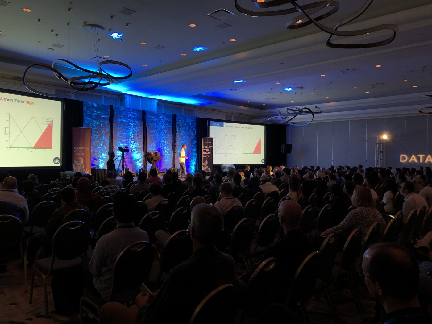 The DataScienceGO ballroom from the back of the room during the session, rows of seated attendees facing two screens that both carry a fuzzy logic slide reading If Quality is Good, then Tip is High.