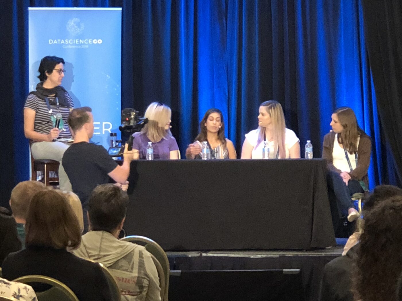 A five-person panel seated at a draped table on the DataScienceGO main stage, a moderator with a microphone on a stool beside a conference banner, audience and camera operator in the foreground.