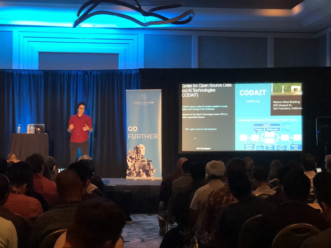 A DataScienceGO session in progress seen from the back of a darkened ballroom, the screen carrying slides on IBM's Center for Open Source Data and AI Technologies.