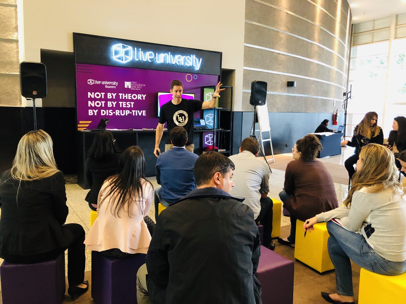 A lobby session at Hotel Caesar Business on 11 June: delegates seated on yellow and purple stools while a speaker presents in front of a Live University Ibramerc screen reading 'not by theory, not by test, by dis-rup-tive'.