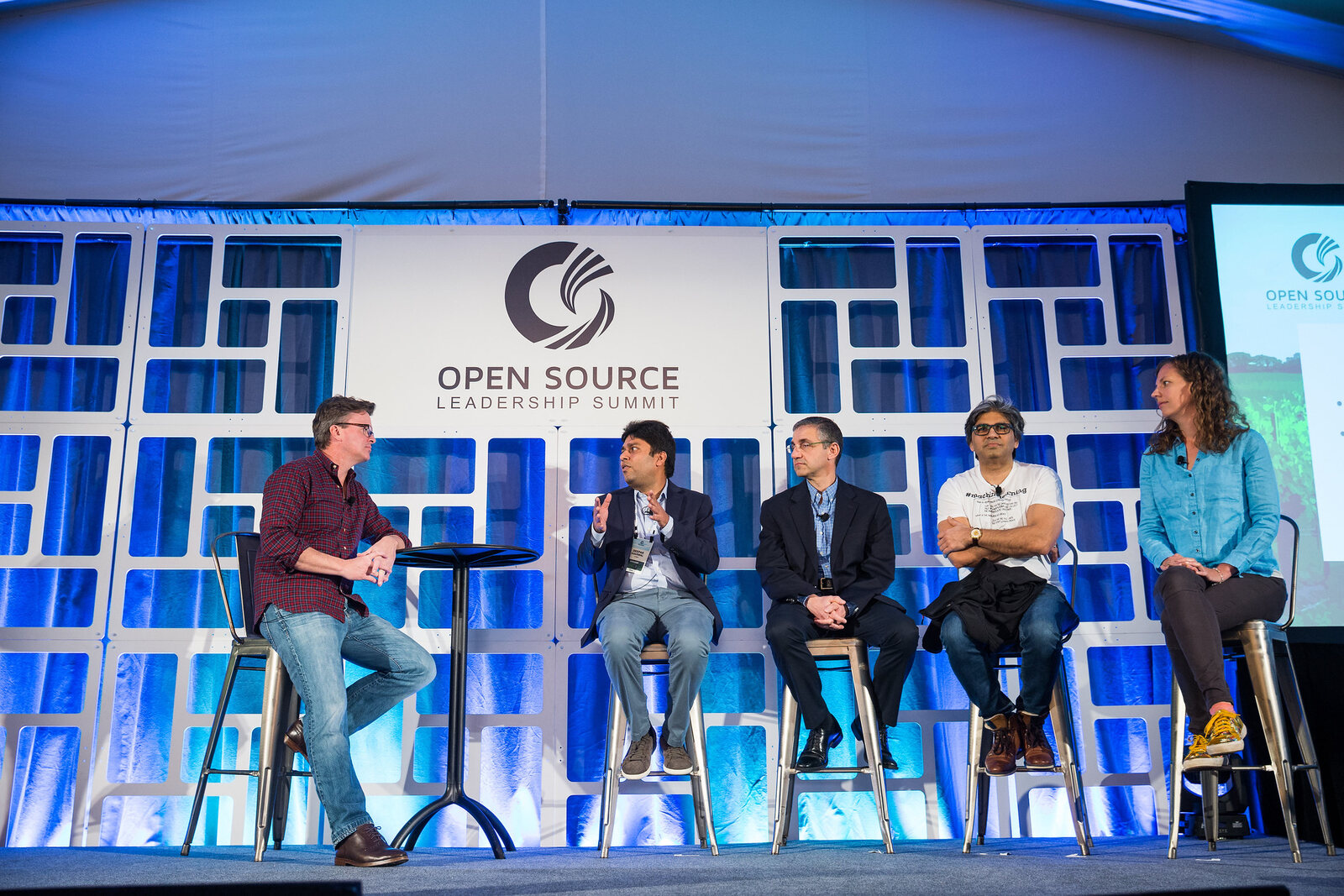 The panel on stage at the Open Source Leadership Summit, a moderator and four panellists on stools in front of the summit's blue backdrop. Tarry Singh sits second from the right in a white #machinelearning T-shirt.