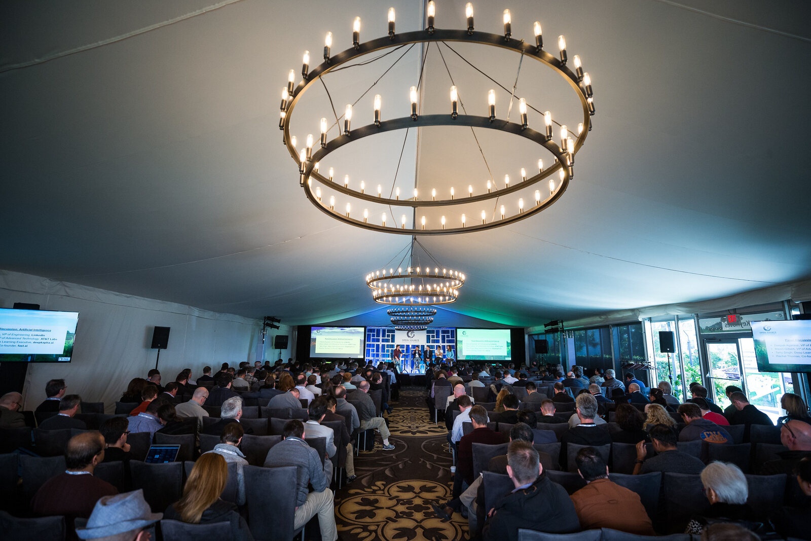The marquee from the back during a session, ring chandeliers down the length of the ceiling and the audience seated in rows towards the lit stage.