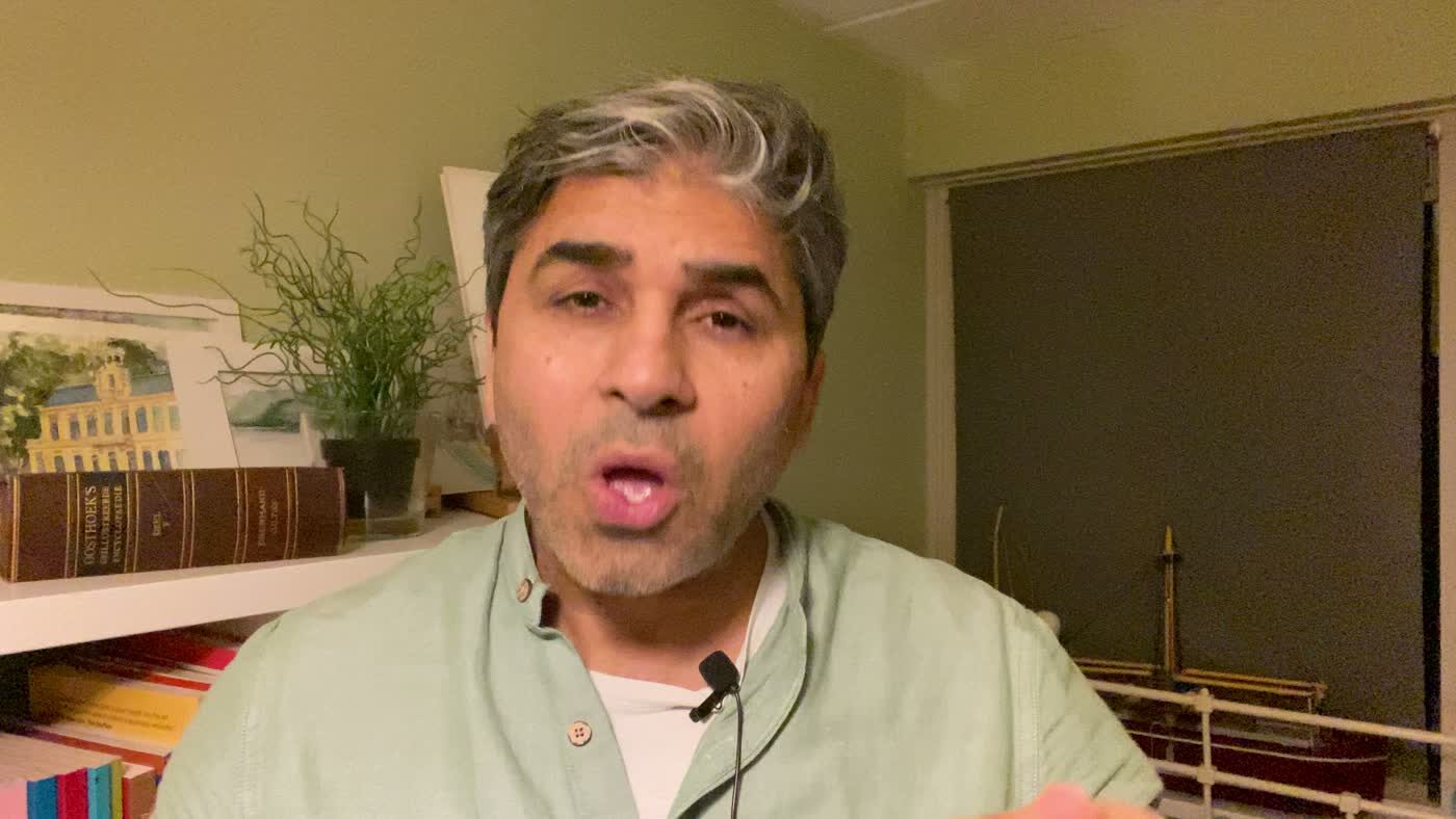 Tarry Singh mid-sentence to camera in a home study, lavalier mic clipped to a pale green shirt, bookshelf and potted plant behind him.