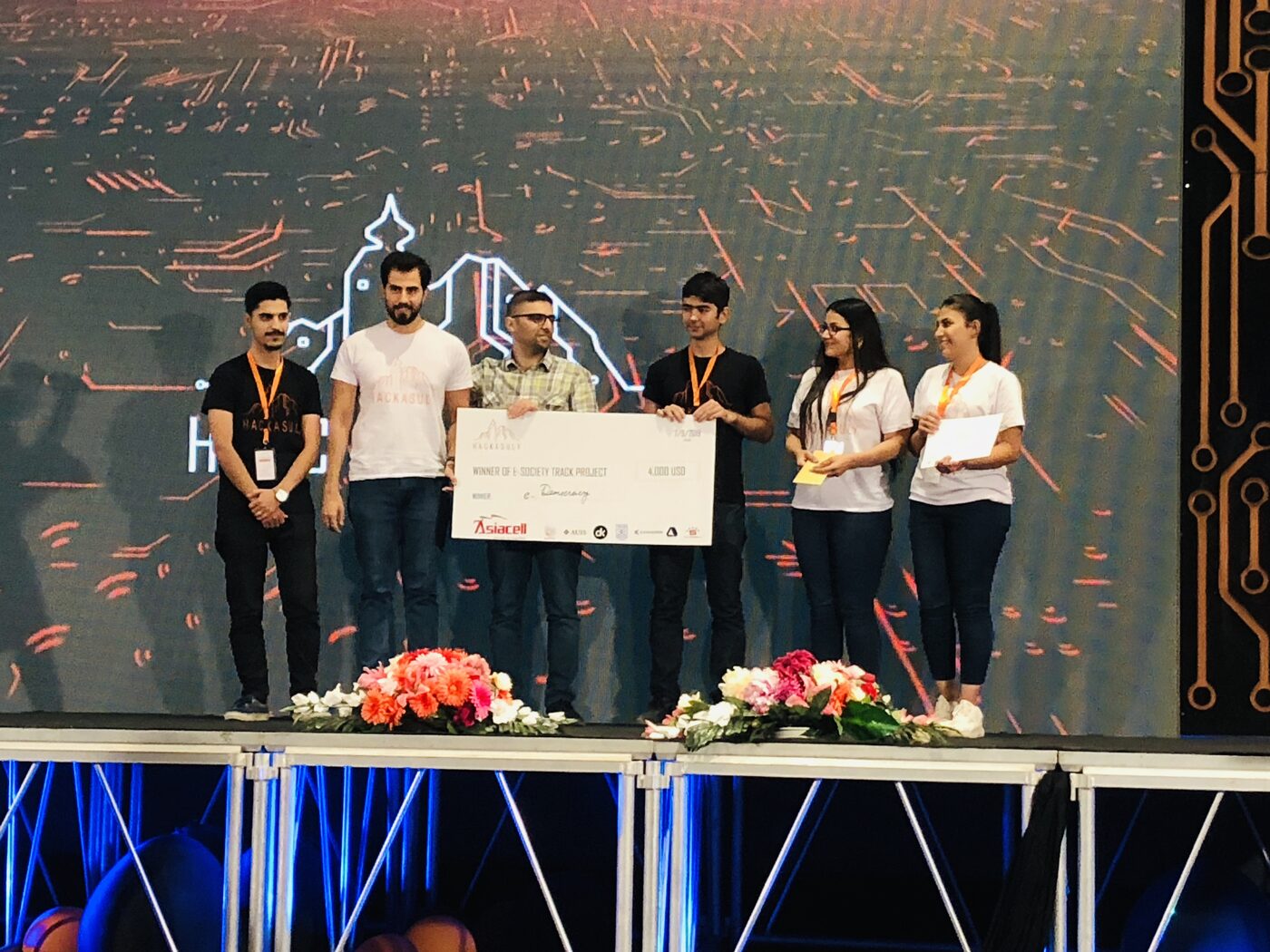 Six hackathon winners stand on the Hackasuly stage holding an oversized cheque reading Winner of e-Society Track Project, 4,000 USD, in front of a circuit-board LED wall.