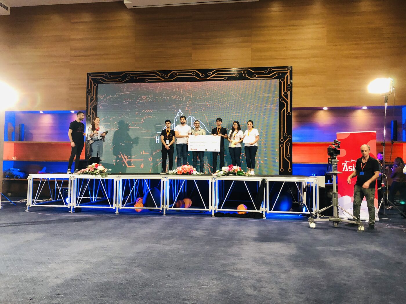 Wide view of the Hackasuly prize ceremony: winners on a low stage with the oversized cheque, a camera operator to the right and an Asiacell banner beside him.