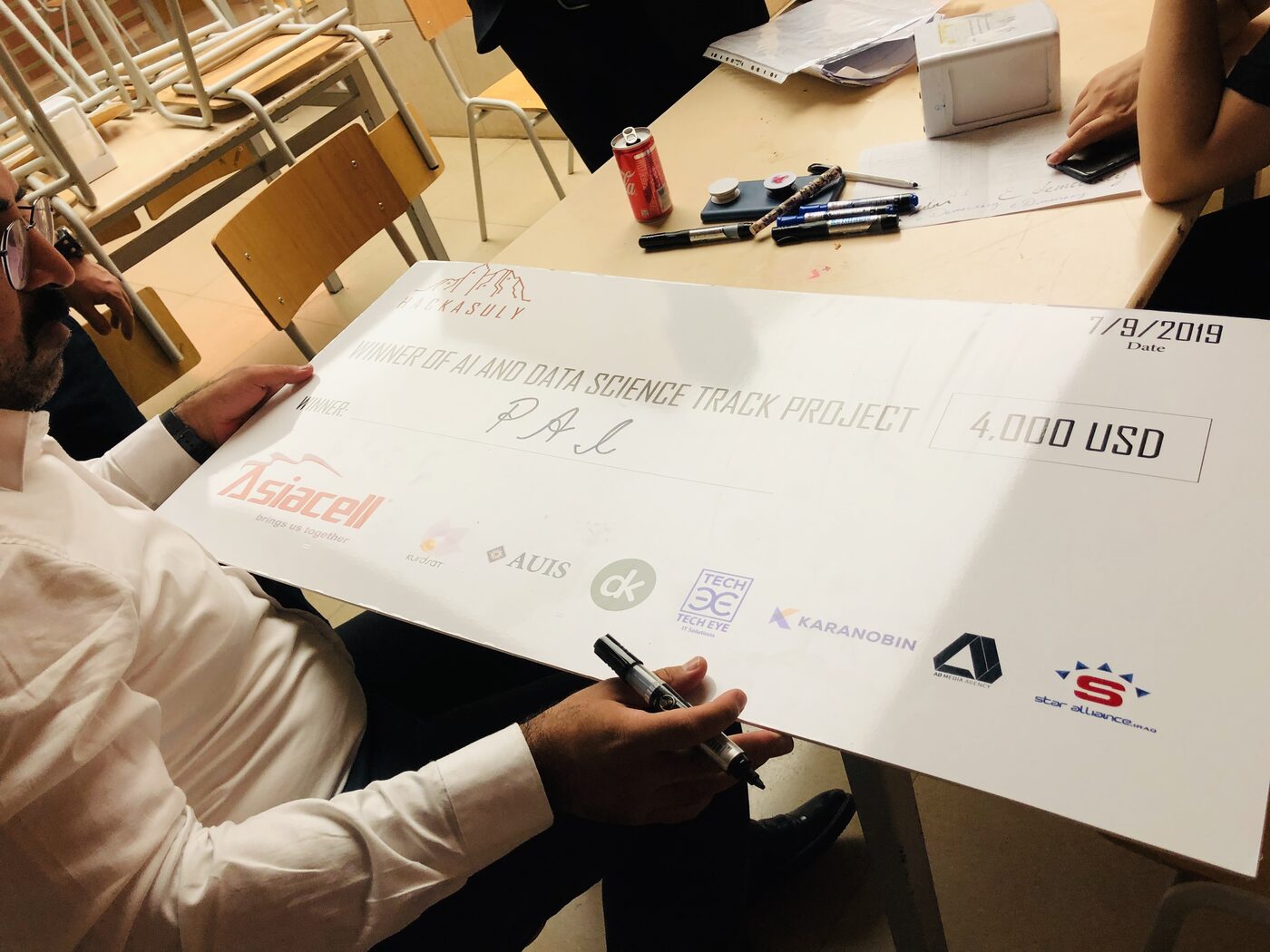 A judge signs the oversized cheque for the AI and data science track, made out to team PAL for 4,000 USD and dated 7/9/2019, with the deepkapha mark in the sponsor row beside Asiacell and AUIS.