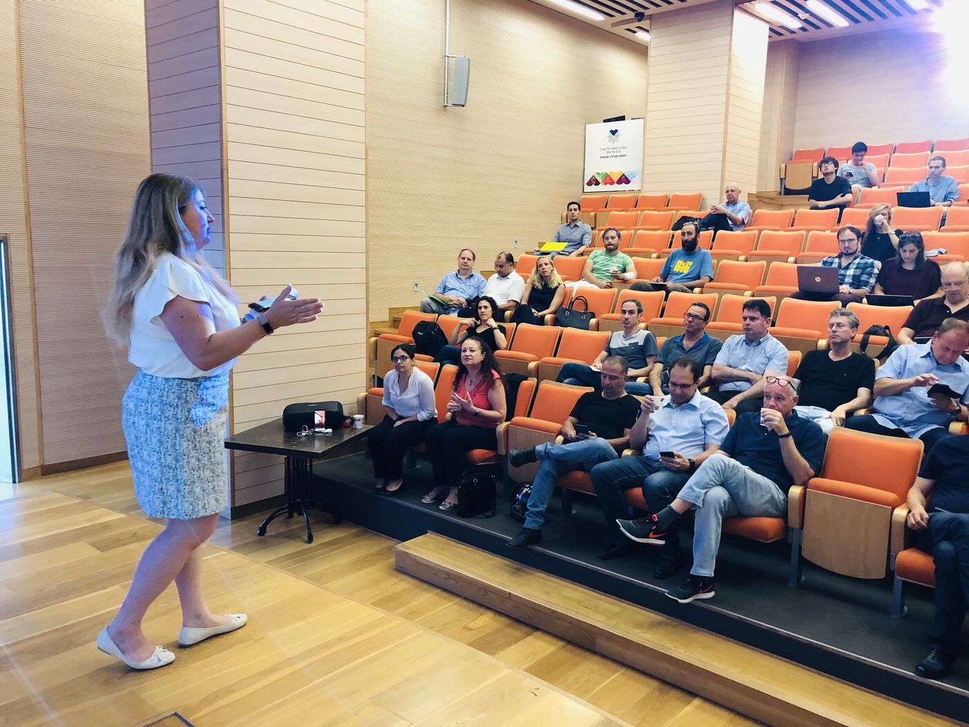 A woman speaks from the floor of the hospital's tiered lecture hall to several dozen seated attendees; the Tel Aviv Sourasky Medical Center sign is mounted on the wall behind the back rows.