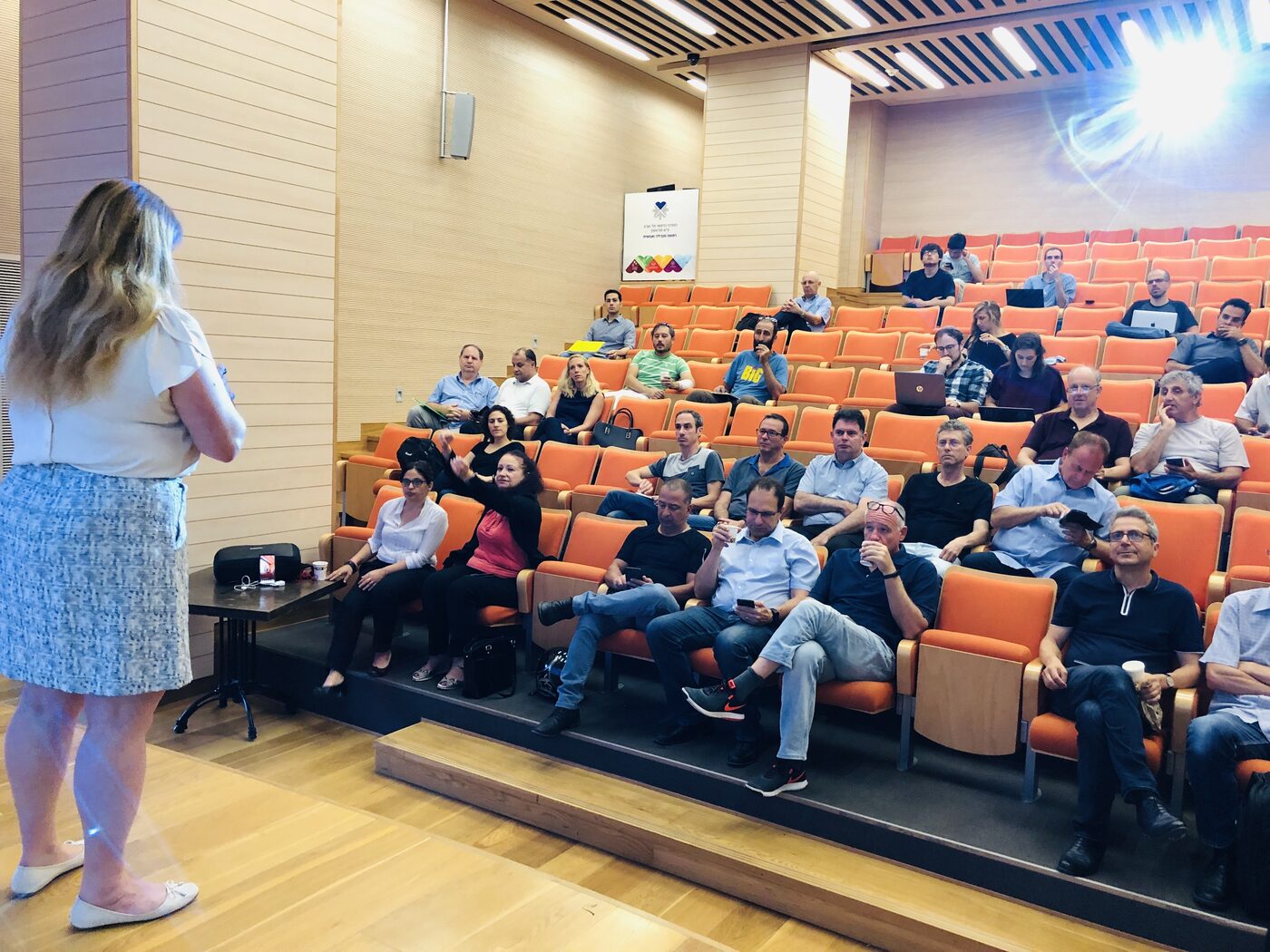 Wider view of the same lecture hall, with attendees across the tiered orange seats, several laptops open on knees.