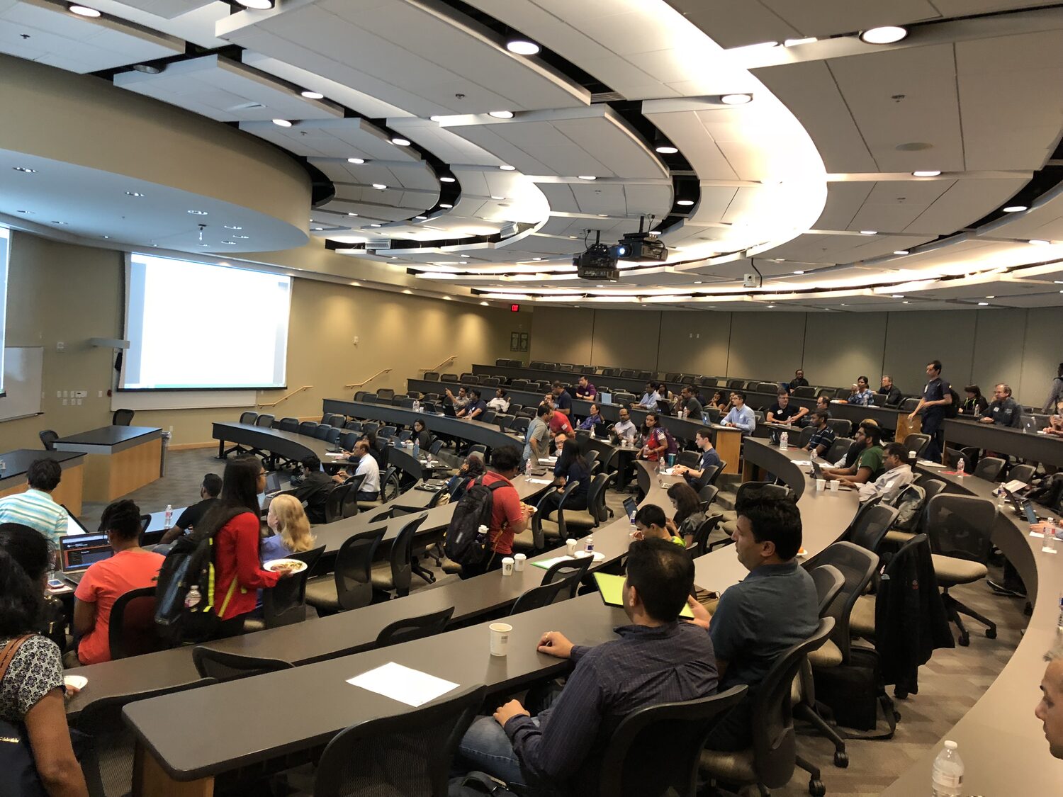 Tiered UT Dallas lecture hall seen from the back, most seats taken, twin projection screens at the front