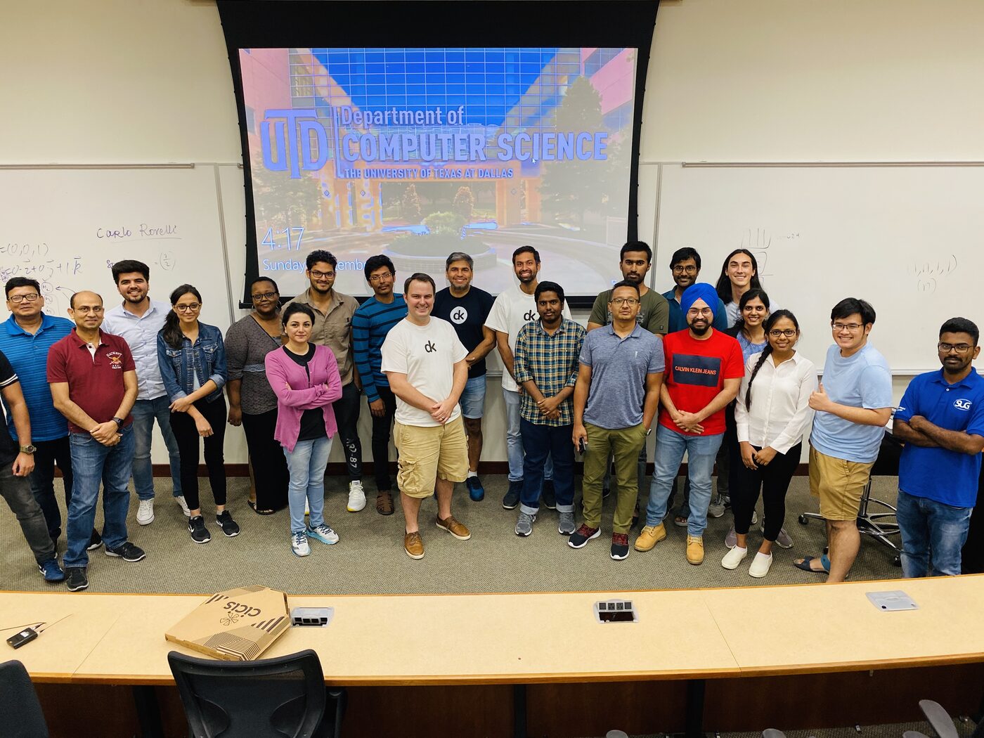 The full cohort and instructors standing together at the front of the UT Dallas lecture hall, with the Department of Computer Science title screen projected behind them.
