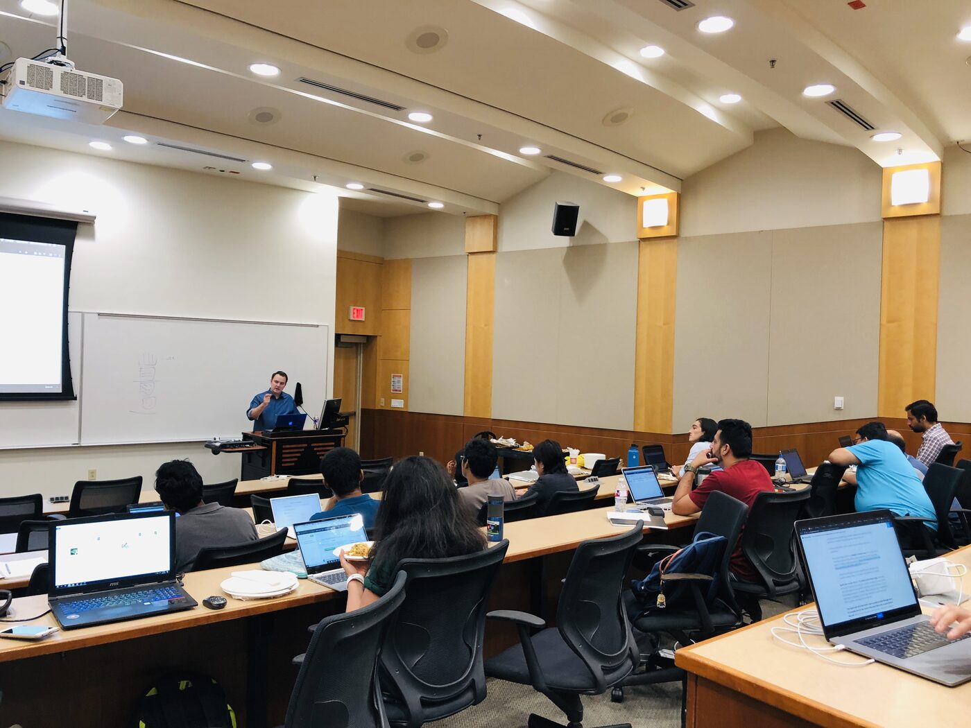 An instructor teaching from the lectern to rows of participants working on open laptops in the tiered lecture hall.