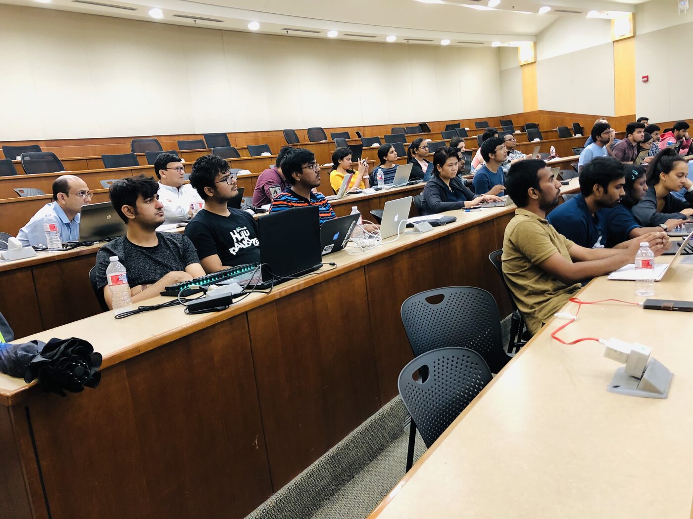 Participants seated along the curved desks of the lecture hall, laptops open, watching the front of the room.
