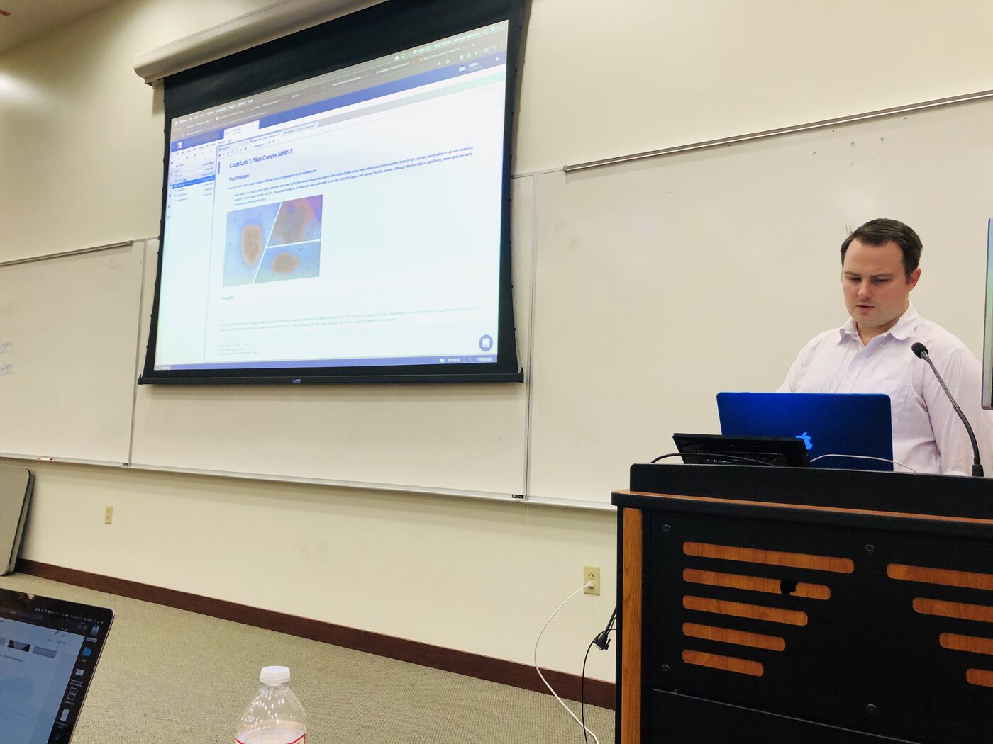 An instructor at the lectern with Code Lab 1: Skin Cancer MNIST projected on the screen beside him.
