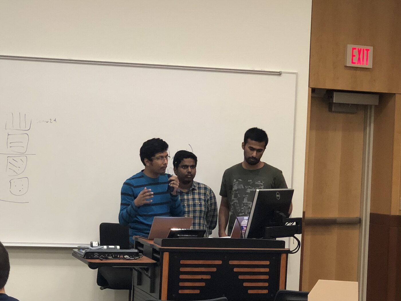 Three team members presenting at the lectern with a handheld microphone on hackathon day.