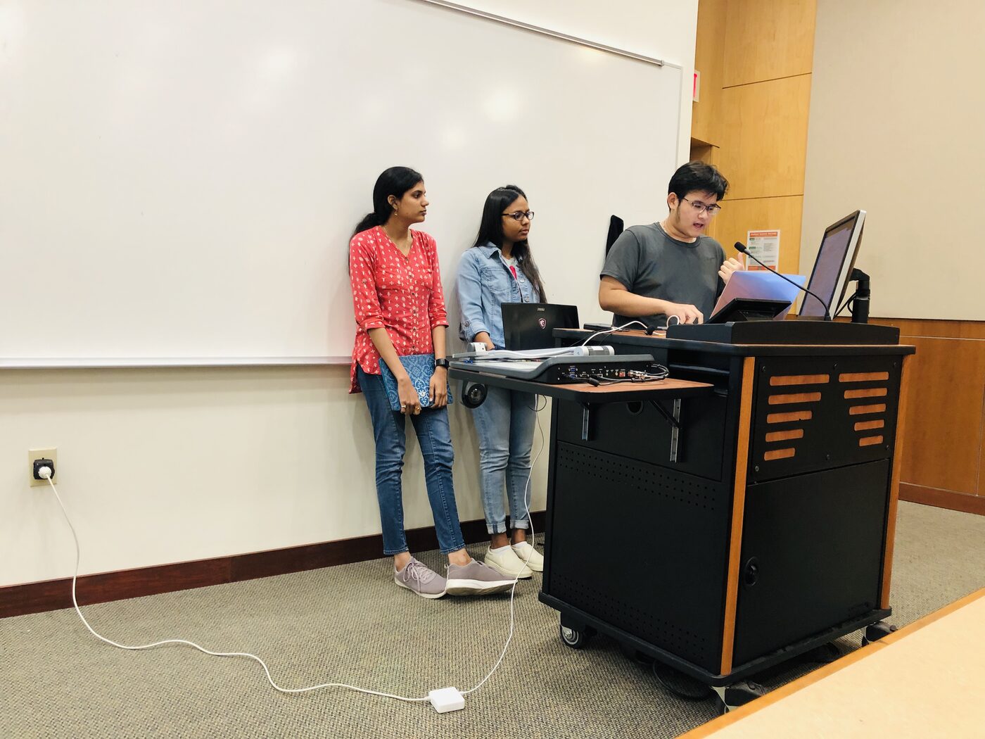 A team of three at the front of the room, one member driving the lectern machine while two stand alongside.