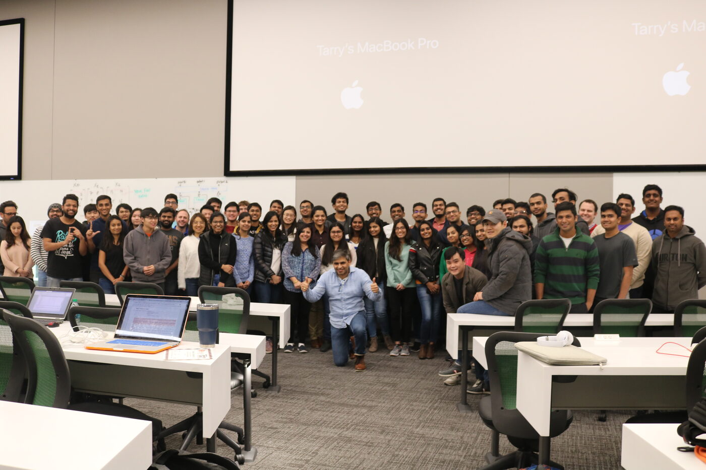 The full cohort standing together across the front of the lecture room on the last day, benches and open laptops in the foreground, whiteboards and projection screens behind them.