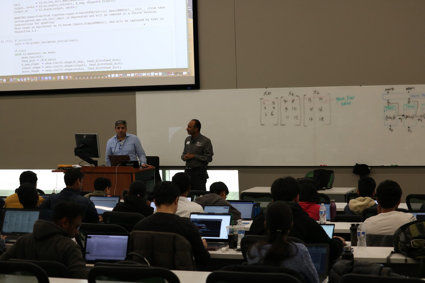 Wide view from the back of the hall: two instructors at the front podium, Python code projected on the screen above them, students at laptops along every bench.