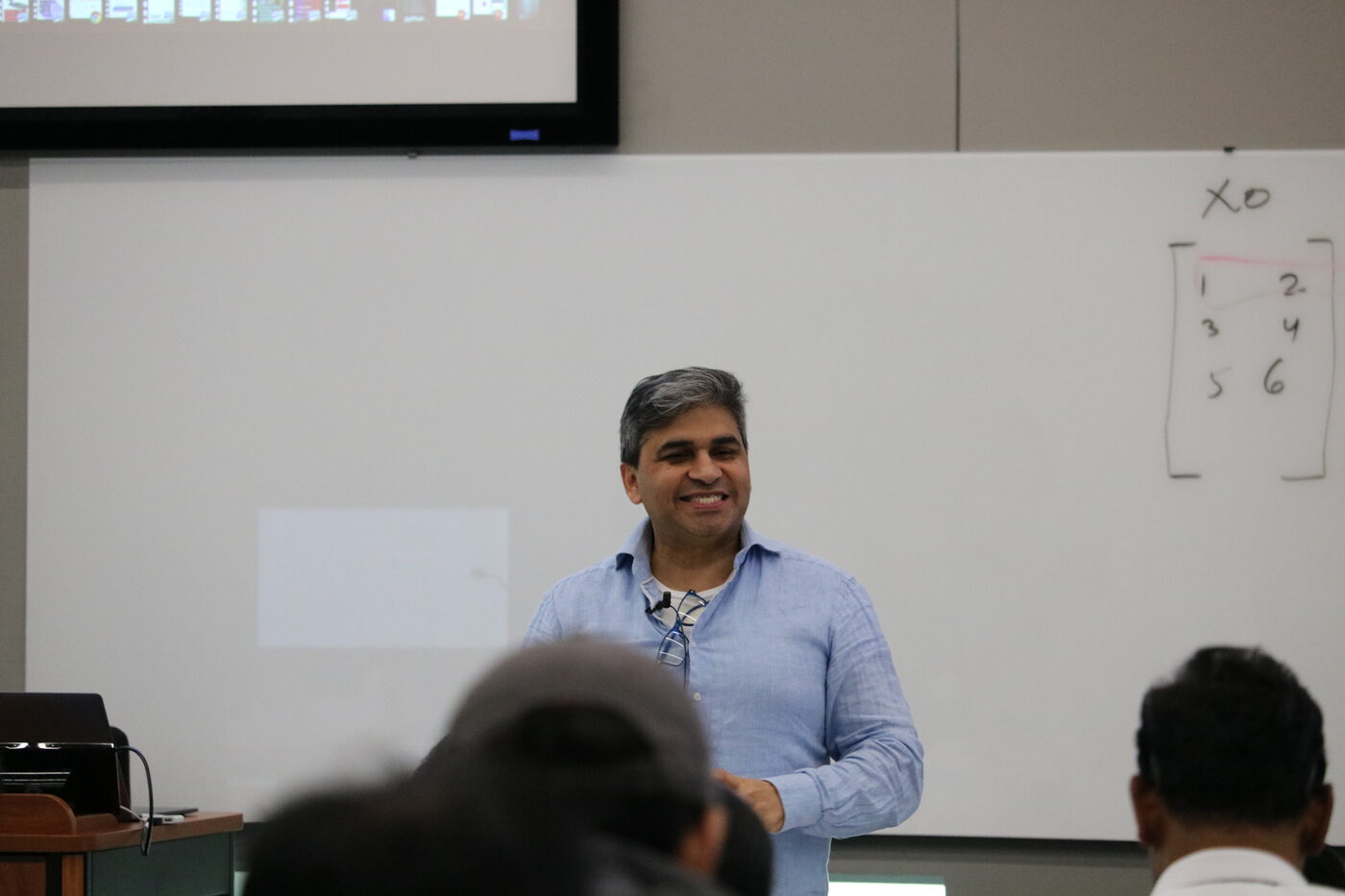 Tarry Singh speaking at the front of the room, a matrix written on the whiteboard behind him, heads of the audience in the foreground.