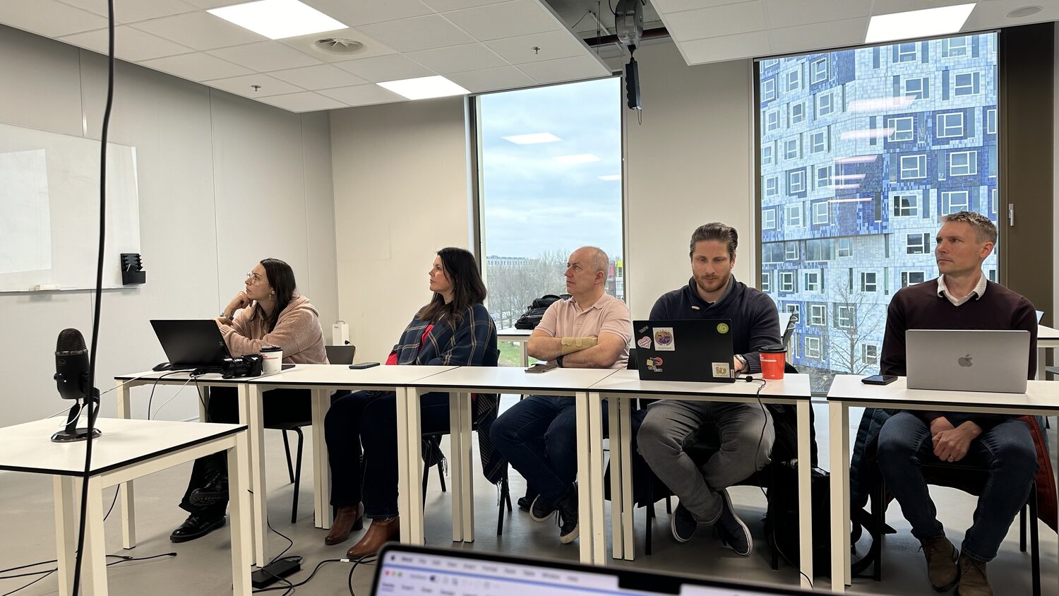 Participants at the HCAIM working session sit along white tables with laptops open, tall windows onto Utrecht Science Park behind them, 31 January 2024.