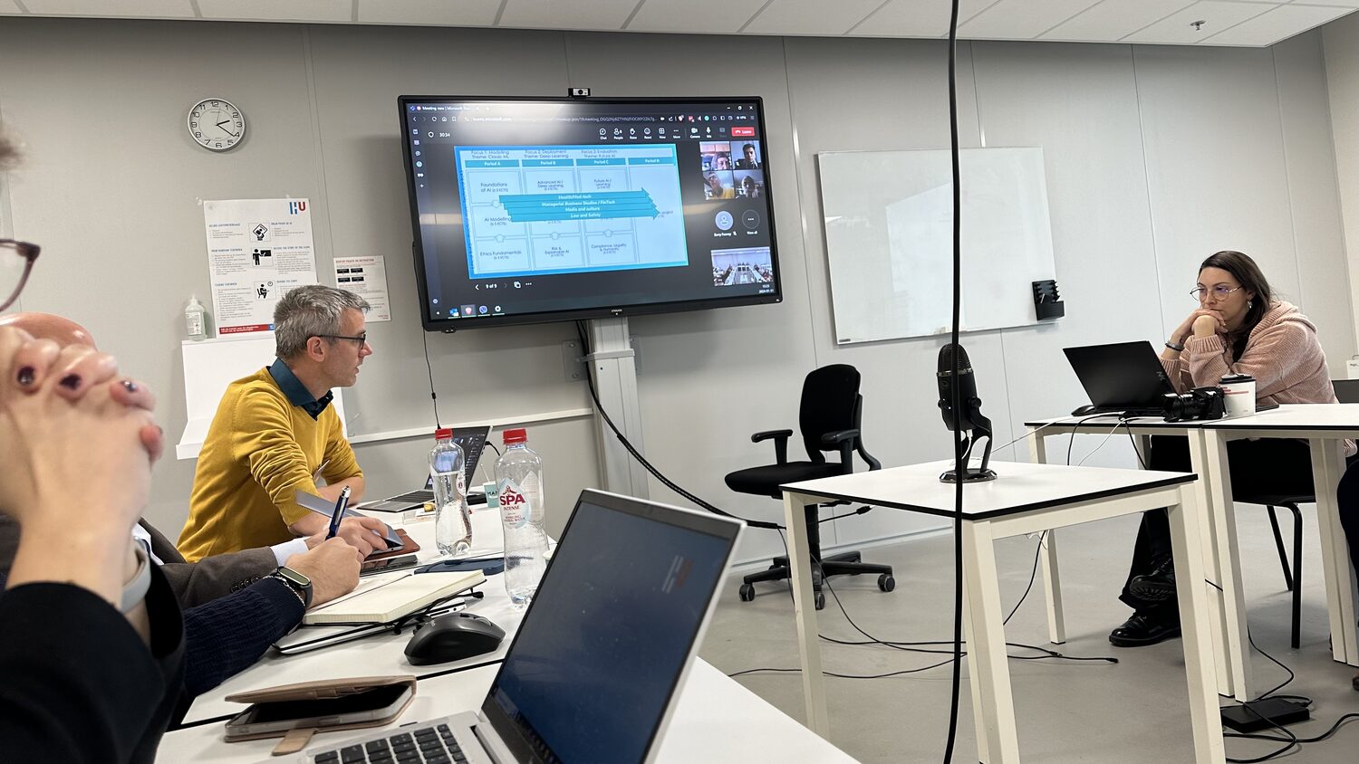 A wall screen shows the HCAIM programme map beside a Microsoft Teams call grid while a participant takes notes in the room, 31 January 2024.