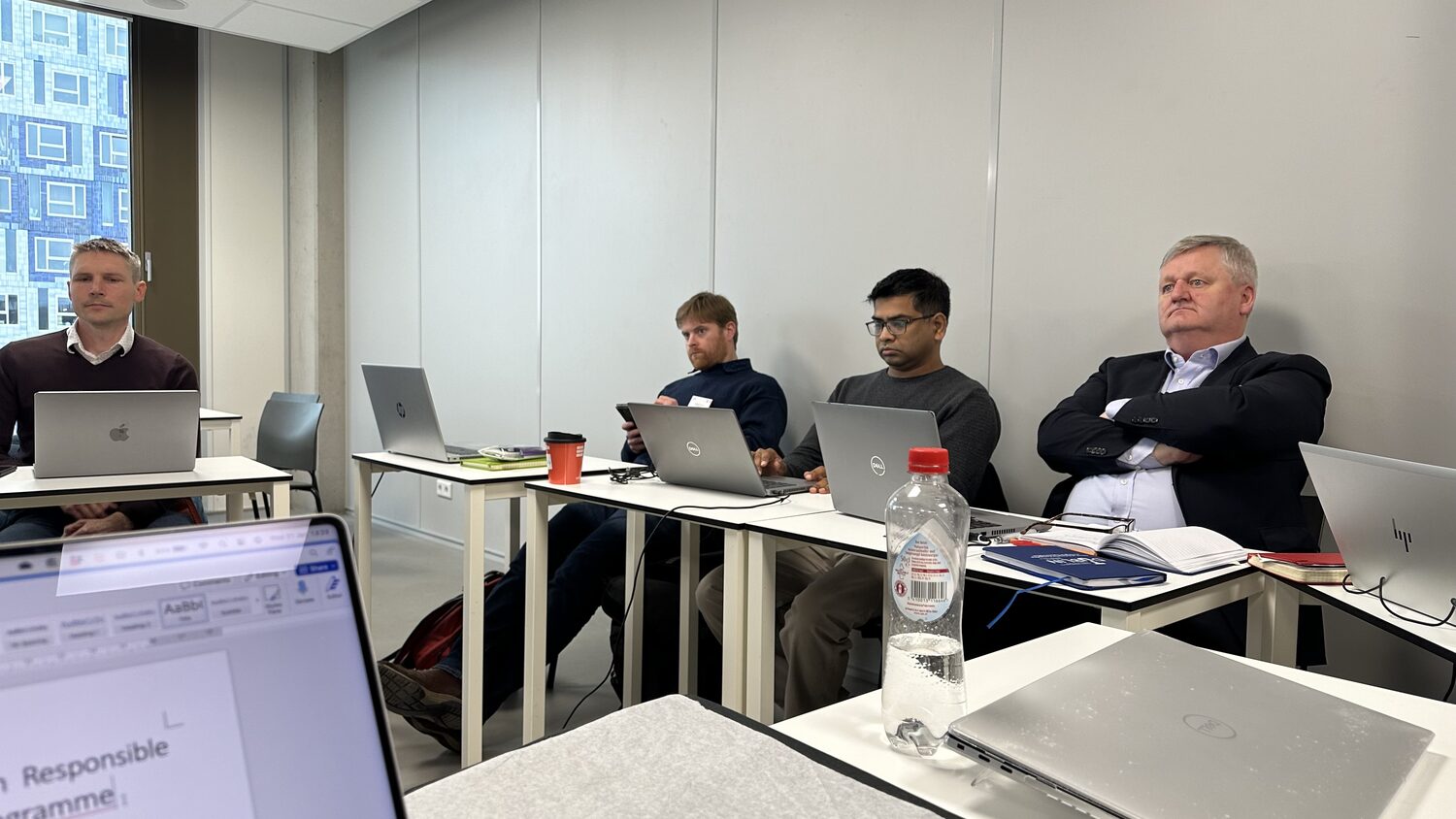 Four seated participants listen during the HCAIM session, laptops, notebooks and coffee cups on the tables.