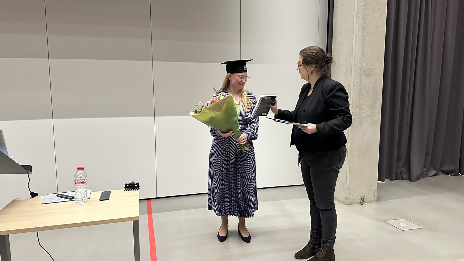 A graduate in cap and gown holds a bouquet while a staff member hands over a bound thesis, in a room at Utrecht Science Park on 31 January 2024.