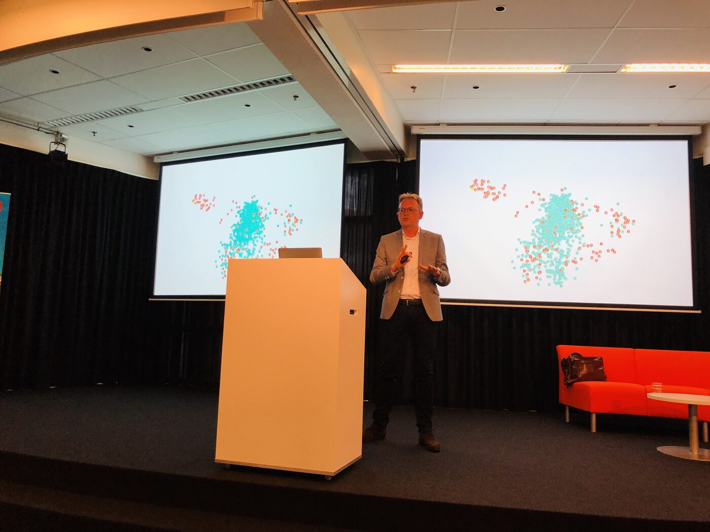 Wide view of the stage: a speaker stands between two projection screens showing the same two-dimensional cluster plot, with a white lectern in front of him and a red sofa at the side of the platform.