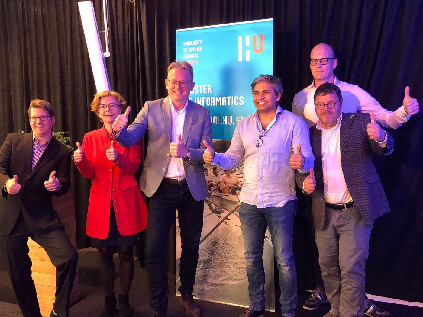 Six speakers and hosts stand in front of the Hogeschool Utrecht Master Informatics banner at the end of the evening.