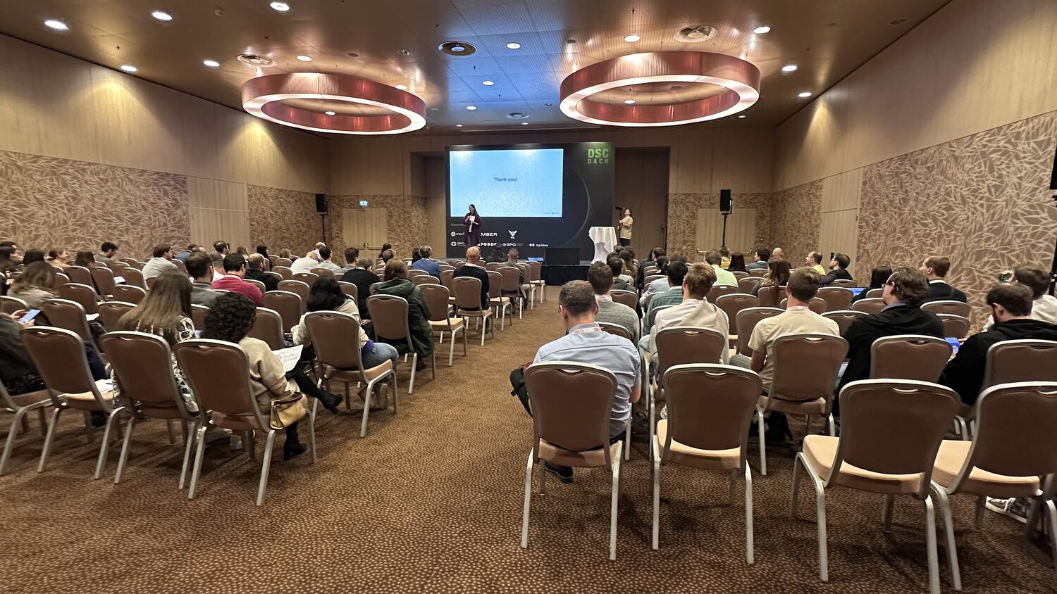 Wide view of the conference room during a session, audience seated in rows, a speaker at the front and the DSC DACH banner beside the screen