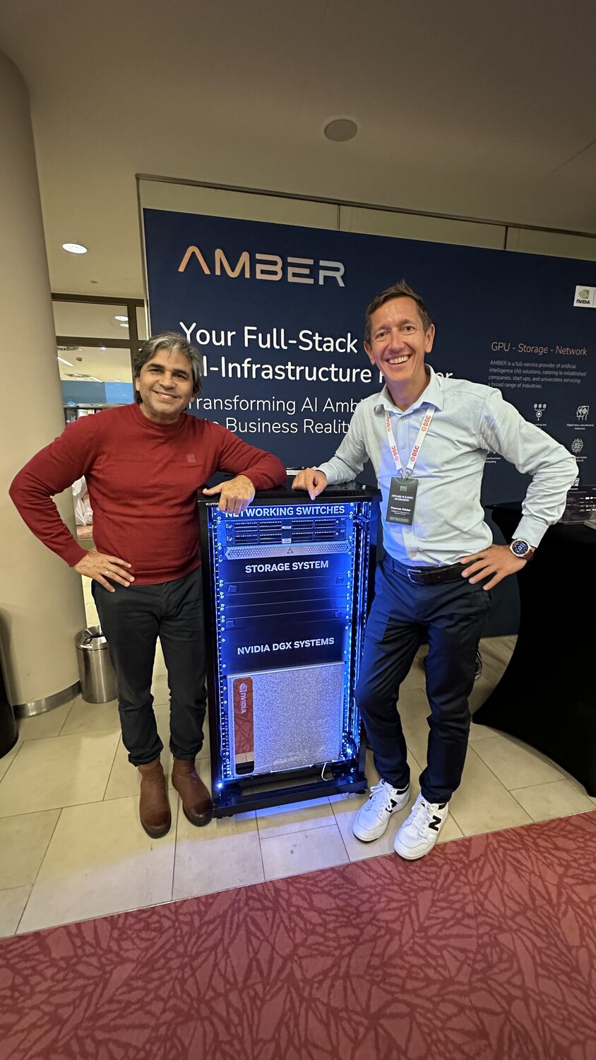 Tarry Singh and a colleague beside a blue-lit GPU rack at the AMBER stand, under a full-stack AI infrastructure banner