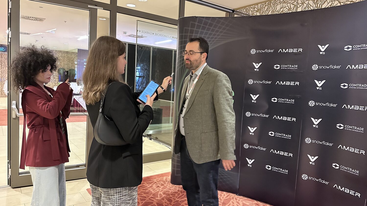 A delegate being interviewed on camera in front of the sponsor wall while a colleague films on a phone