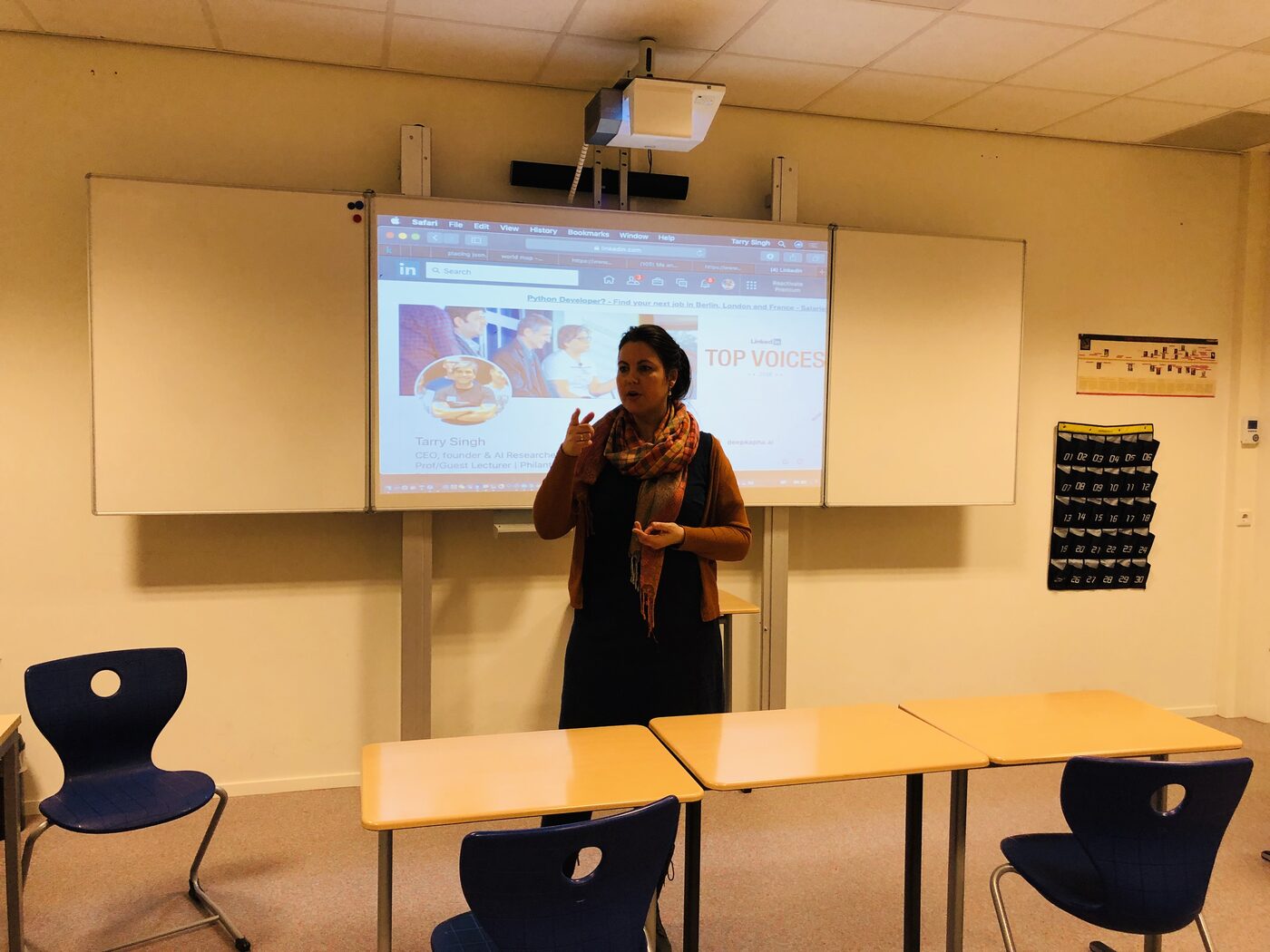 Wide view of the classroom: Foke Van Der Helm stands centre-frame in front of the whiteboard with Tarry Singh's LinkedIn profile projected behind her, empty desks and chairs in the foreground.