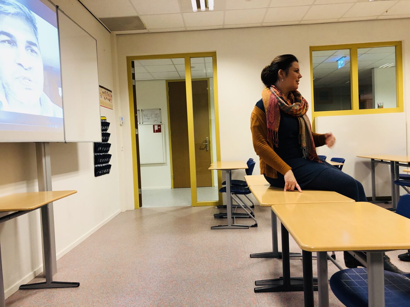 The classroom seen from the back: the speaker perched on the edge of a desk while a video plays on the projection screen at the far left.